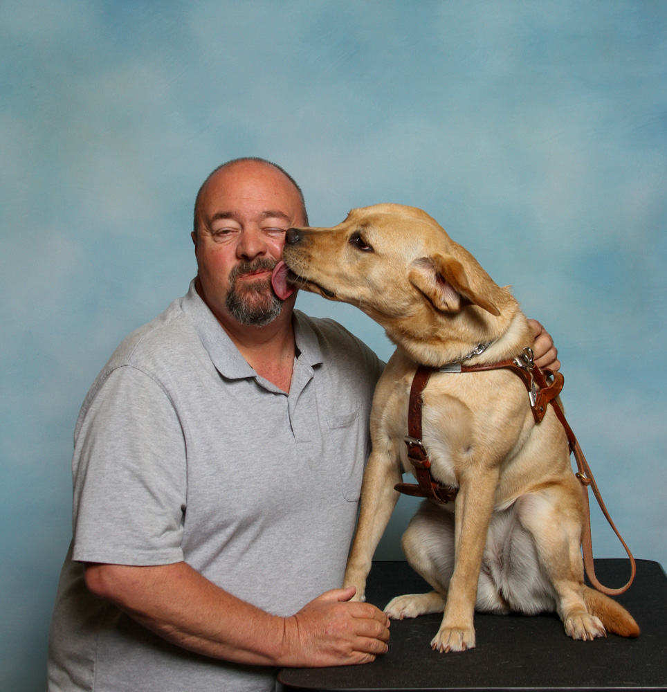 Seeing Eye graduate Jeffrey poses for a photo in front of a blueish backdrop with his yellow Lab Seeing Eye dog, Archie, for their new portrait. Rather than look straight towards the camera, Archie turns and licks the face of Jeffrey.