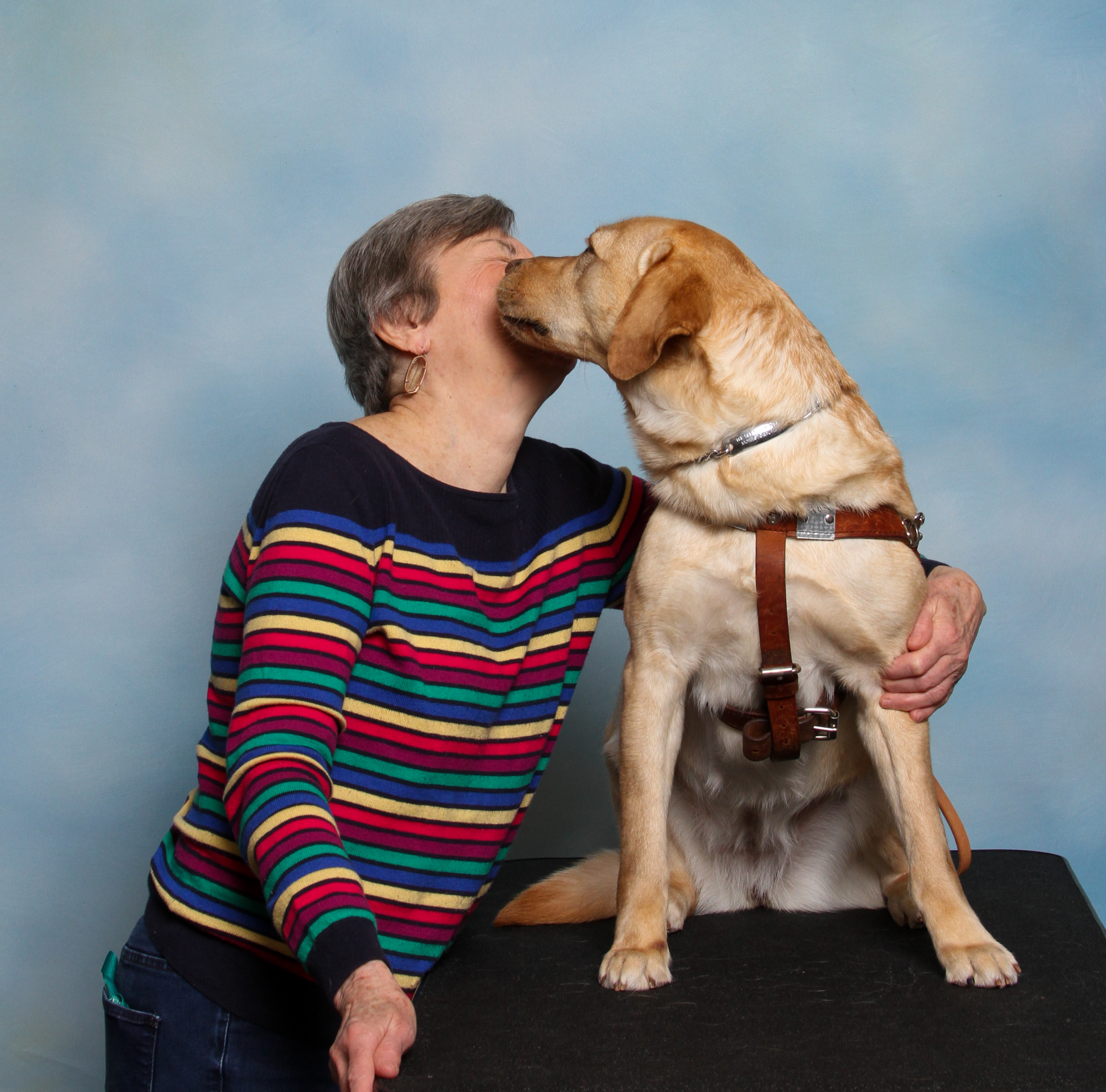 Seeing Eye graduate Susan poses for a portrait with her new yellow Lab Seeing Eye dog Goldy. Susan’s head is tilted back and up as Goldy turns to lick her face.