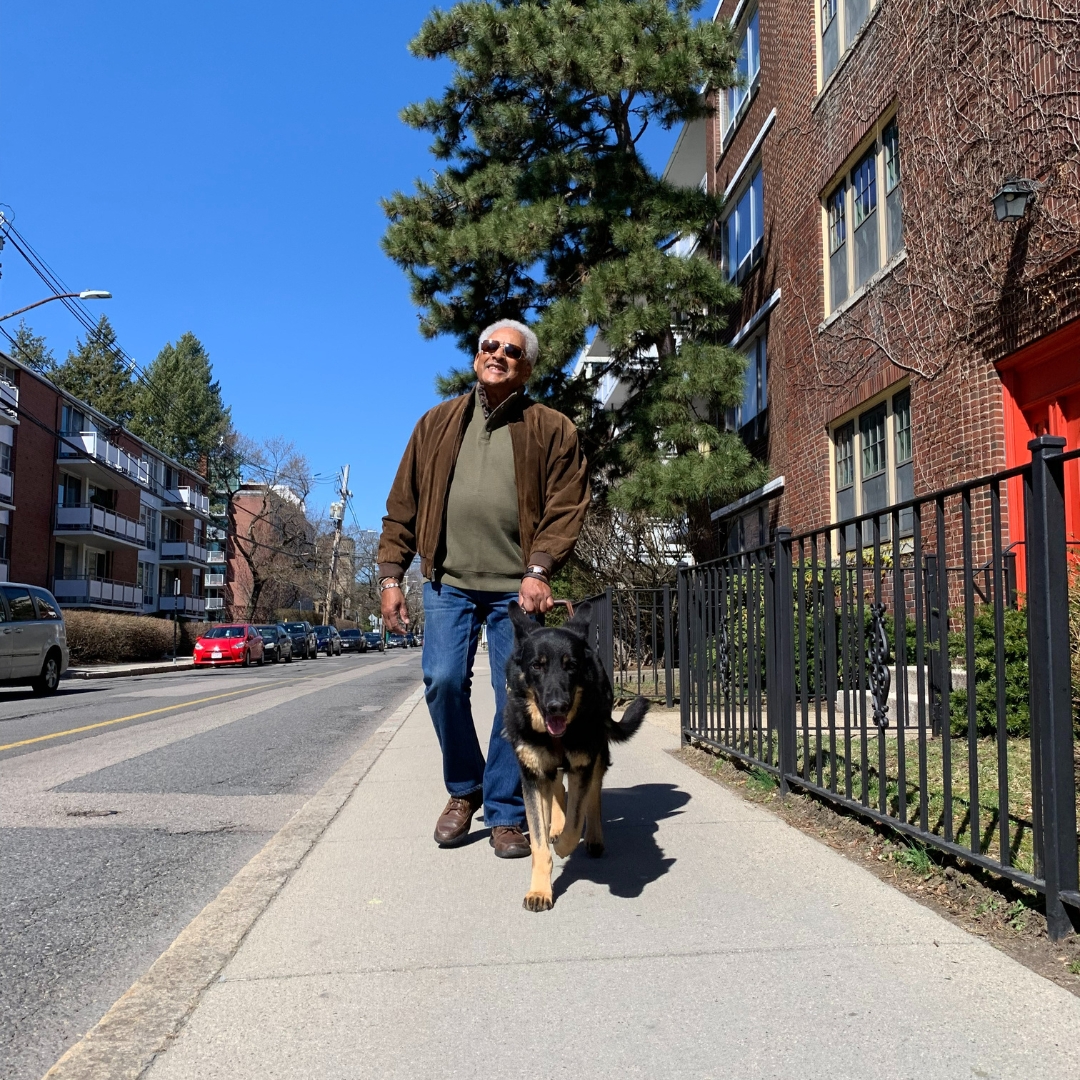 Dean is guided by Gus, a black and tan German shepherd, along the sidewalk of an apartment building.