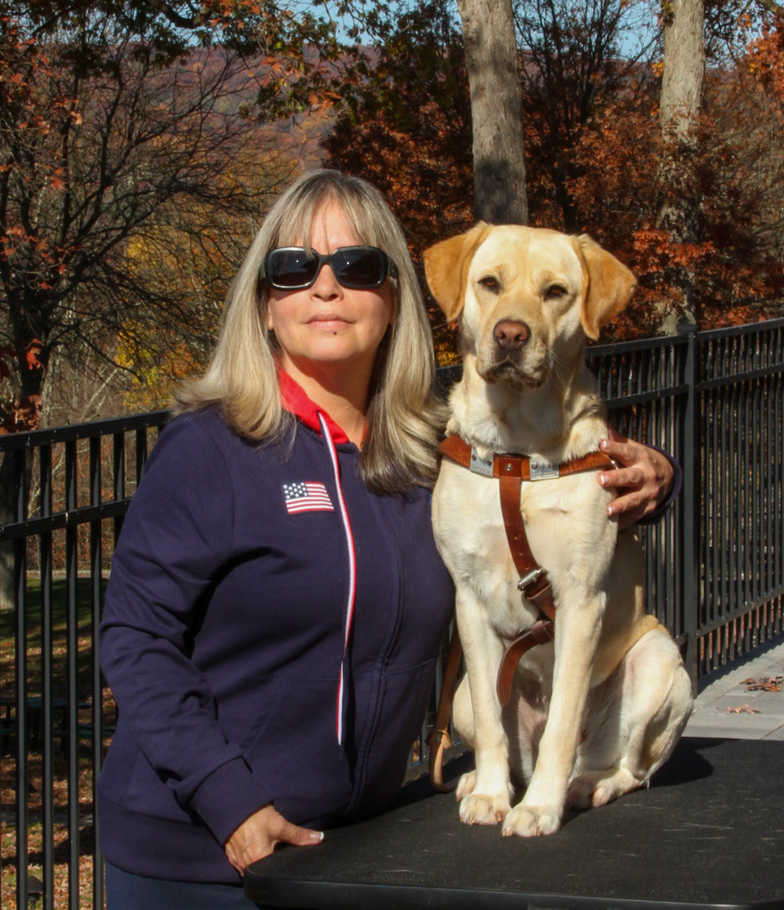 Luz stands with her arm around Verna, a yellow Lab/golden cross in harness, who sits on a raised platform in front of trees full of fall foliage.