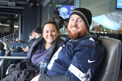 A young woman and a bearded man are smiling as they sit in leather-backed stadium seats. The glass windows of the luxury suite are visible behind them.