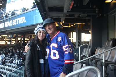 A young woman and man stand near their stadium seats. They are smiling. The woman has a Cowboys hat and the man has a Giants jersey.