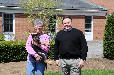 Cheryl and Glenn stand outside, smiling next to each other. Behind them, is the brick facade of the main building at The Seeing Eye campus. Cheryl is holding a small German shepherd puppy in her arms.