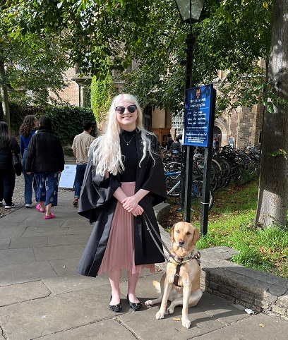 Sarah stands in a black graduation gown with her yellow Lab/golden cross Seeing Eye dog, Elana, sitting by her side in front of the Trinity College Chapel at the University of Cambridge.