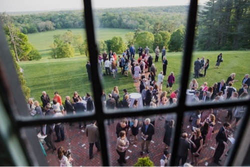 An image from one of the upper floors of the mansion at Natirar – Ninety Acres. A crowd of people enjoys the cocktail hour on the patio with a sprawling green landscape in the background. 