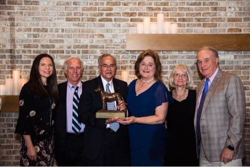 Dr. Gus Aguirre, recipient of the Dorothy Harrison Eustis Award is shown standing against a candlelit wall with his award, a sculpture of Buddy, and surrounded by fellow University of Pennsylvania Allumni. L to R: Seeing Eye Attending Veterinarian Dr. Kristen Polo, Seeing Eye Trustee Dr. Anthony DeCarlo, Dr. Gus Aguirre, Seeing Eye Director of Canine Medicine and Surgery Dr. Dolores Holle, Seeing Eye Trustee Dr. Vickie Myers-Wallen, Dr. Thomas Trotter. 
