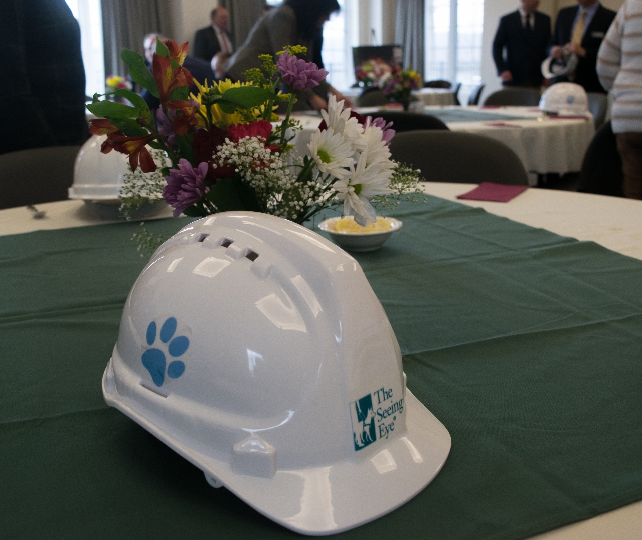 Close up of the white construction hats worn at the groundbreaking ceremony. It has a green Seeing Eye logo on the front and a blue paw print on the side. It's resting on a table at the luncheon held before the ceremony. 