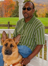 Carlos sits with a black and tan German shepherd under a white gazebo with fall foliage in the background.