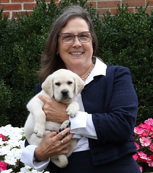 Daphne smiles while holding an 8-week-old yellow Lab puppy. White and pink flowers border green shrubs in the background behind her. 